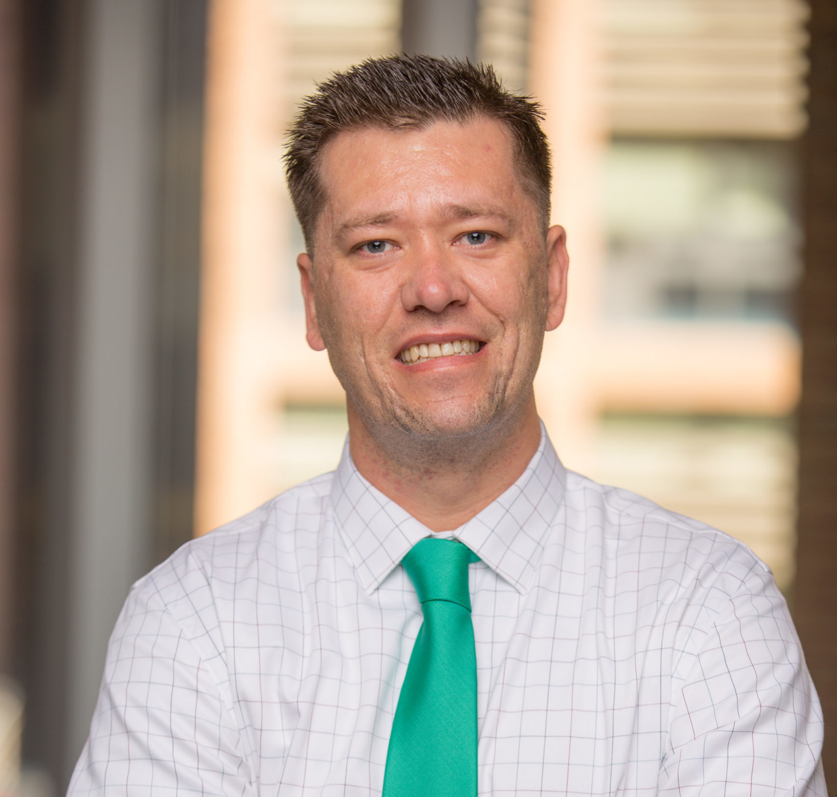 A man with short brown hair smiles at the camera. He is wearing a white checkered shirt and a bright green tie, standing indoors with blurred windows in the background.