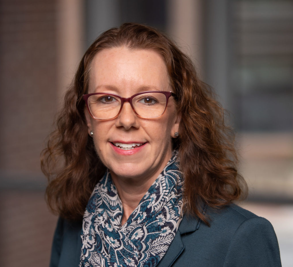 A woman with curly brown hair wearing glasses, a blue patterned scarf, and a dark blazer, smiling in front of a blurred indoor background.
