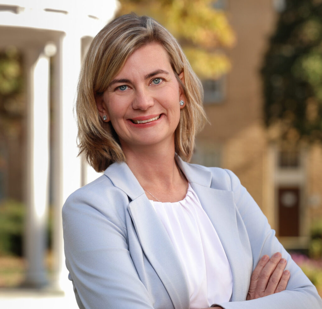 A woman with shoulder-length blonde hair wearing a light blue blazer and white blouse stands outdoors, smiling with her arms crossed. A white columned structure and blurred greenery are visible in the background.