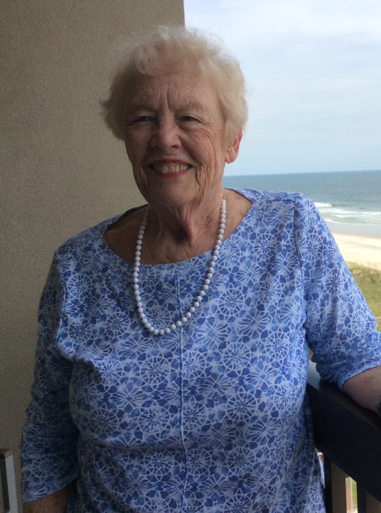 Landy Fox, wearing a blue patterned top and a pearl necklace, smiles while standing on a balcony overlooking a sandy beach and the ocean.