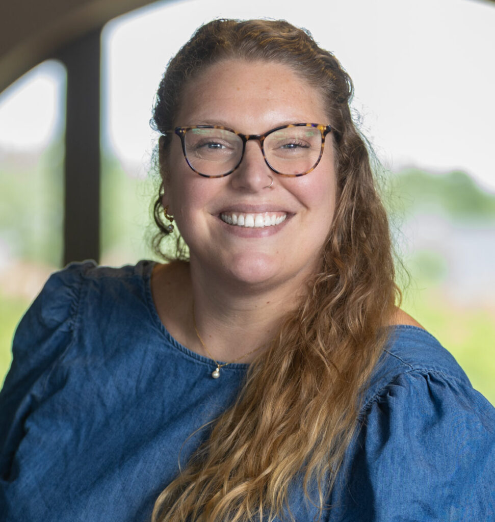 A woman with long, wavy hair and glasses smiles while standing indoors near a window. She is wearing a blue blouse and the background is softly blurred with natural light coming through the window.