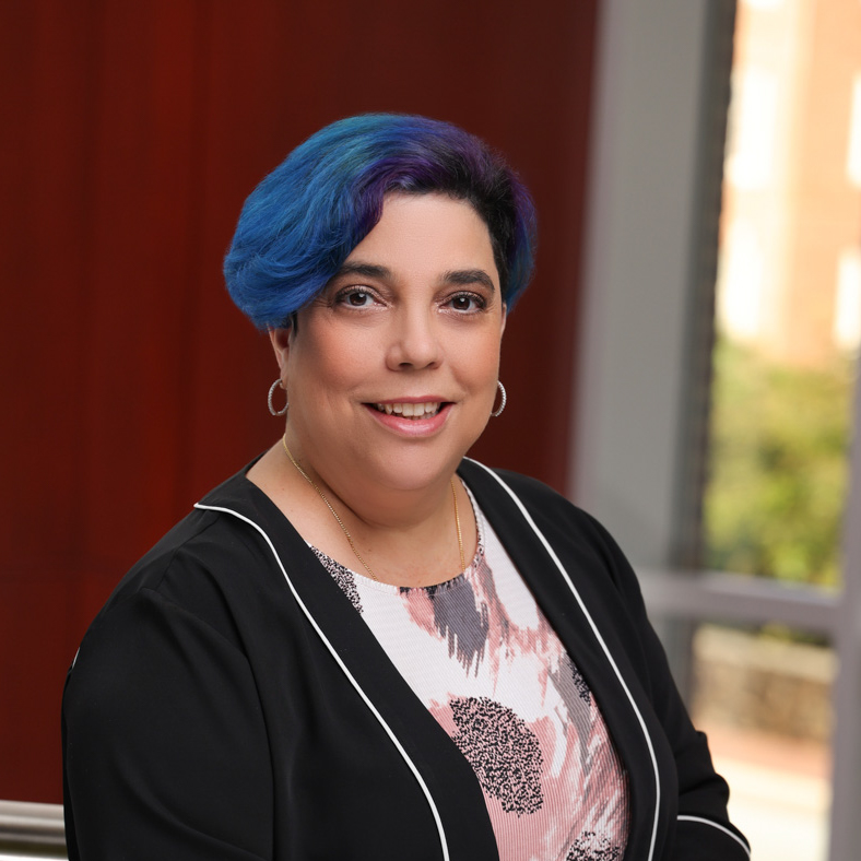 A person with short blue and purple hair, wearing a black jacket with white trim over a floral blouse, smiles while posing indoors in front of a blurred window and reddish-brown wall.
