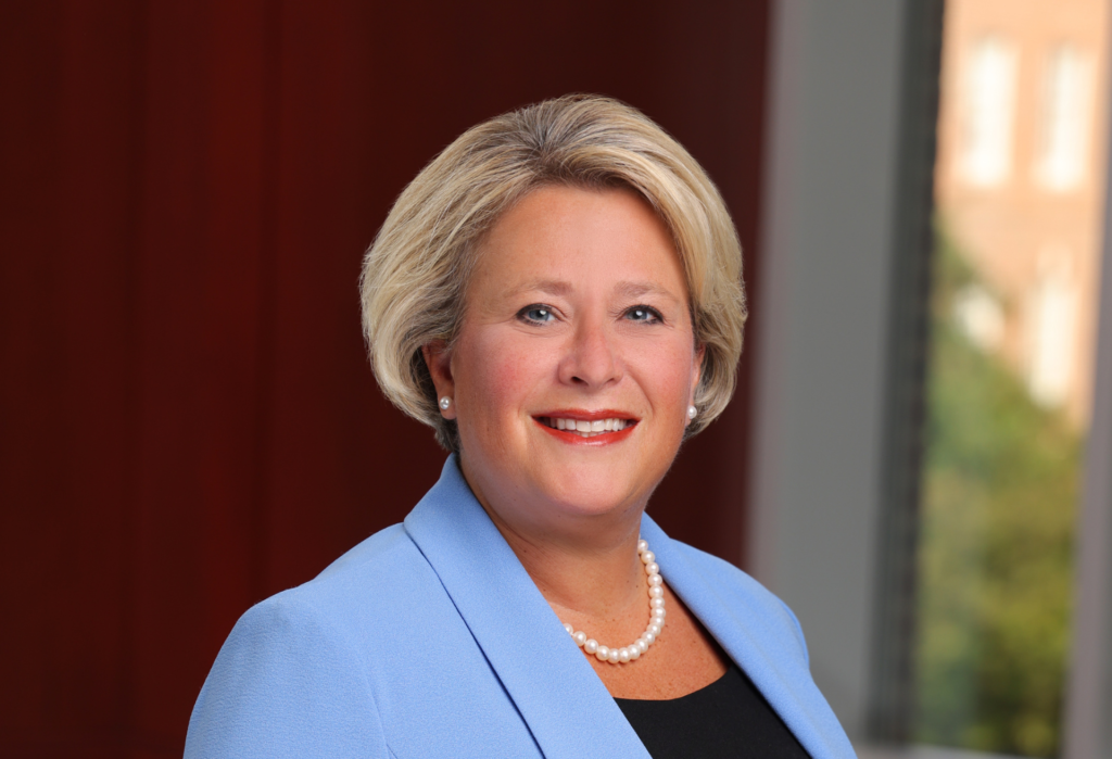 A woman with short blonde hair wearing a light blue blazer, black top, pearl necklace, and pearl earrings smiles in a professional indoor setting with a blurred background.