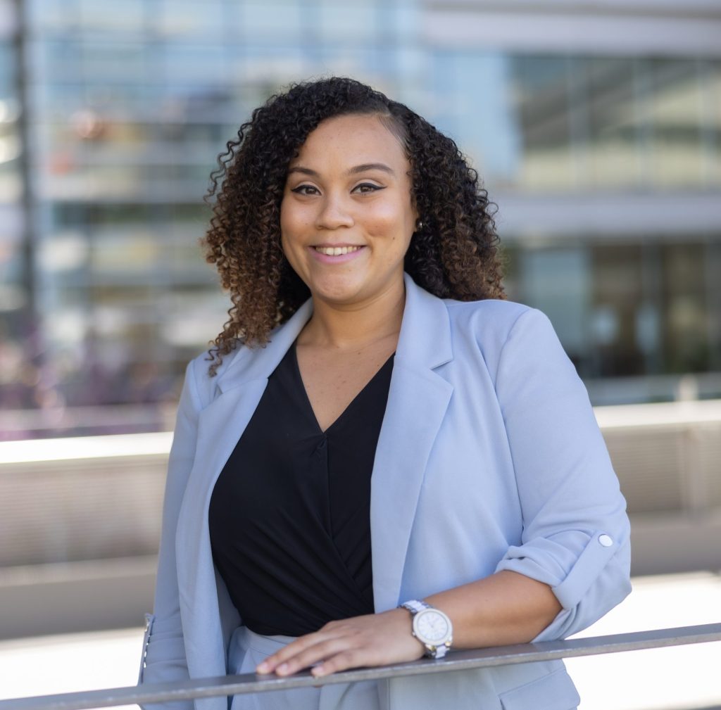 A woman with curly hair, wearing a light blue blazer and black top, stands smiling outdoors in front of a modern glass building.