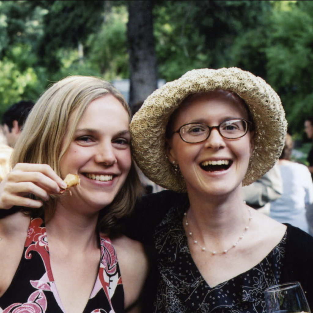 Two smiling women at an outdoor gathering, one holding food and the other wearing glasses and a straw hat. Both appear happy, surrounded by greenery and blurred people in the background.