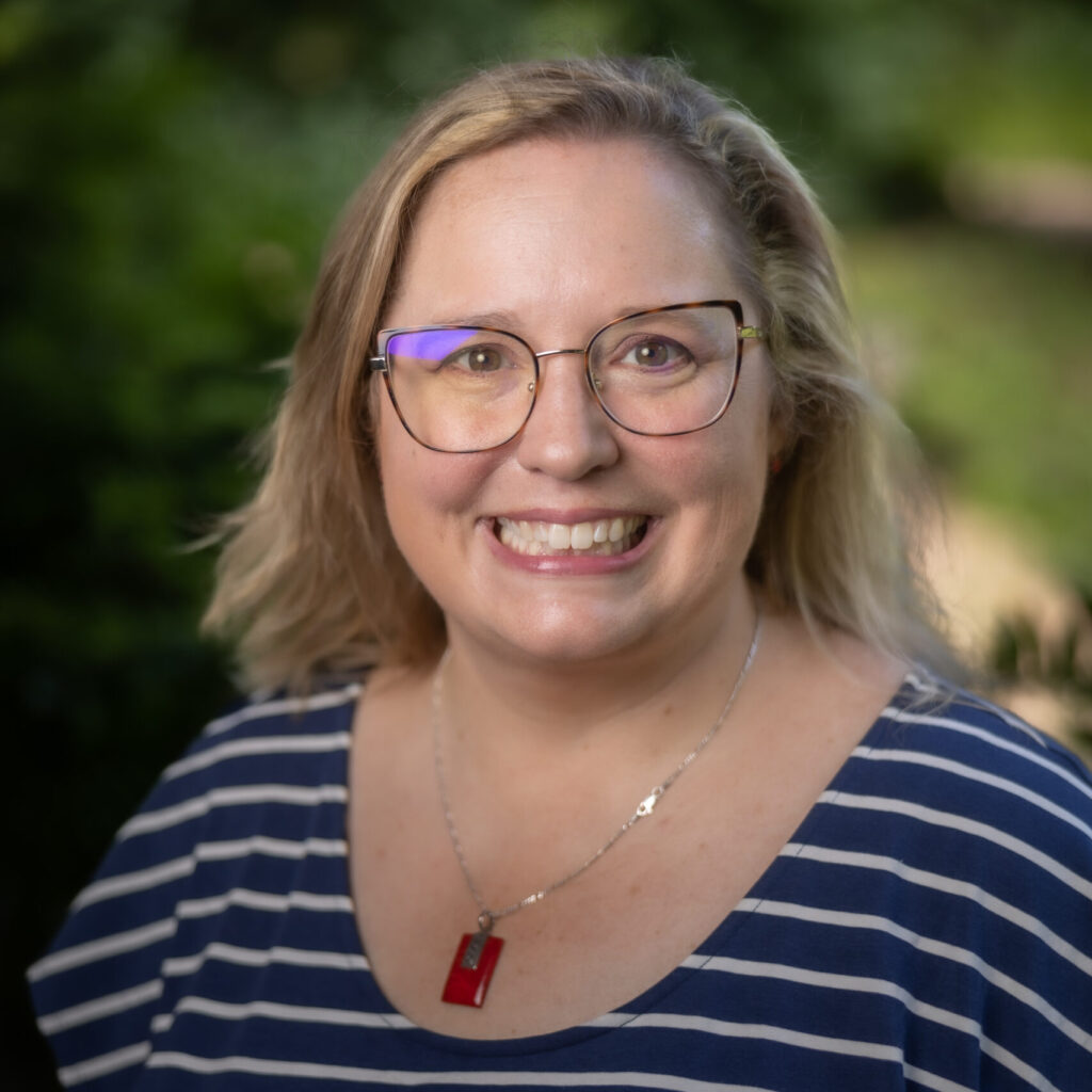 A smiling woman with blonde hair and glasses wears a blue and white striped shirt and a red pendant necklace, standing outdoors with greenery in the blurred background.