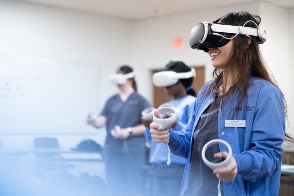 Three nuraing students in a classroom wear virtual reality headsets and hold controllers, actively participating in a VR simulation. The person in the foreground is smiling and wearing blue Carolina Nursing scrubs.