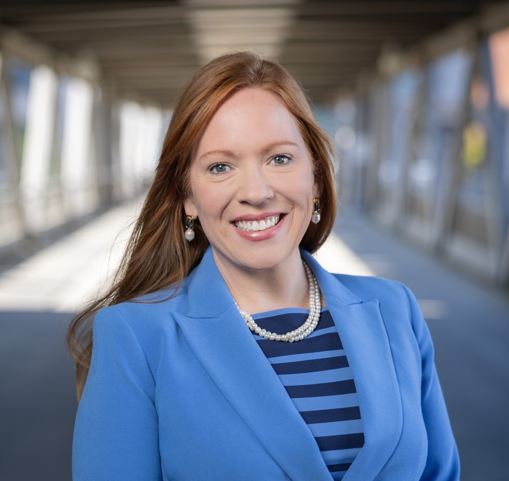 A woman with long red hair smiles, wearing a blue blazer, striped navy shirt, pearl necklace, and earrings, standing in a bright, covered walkway with blurred background.