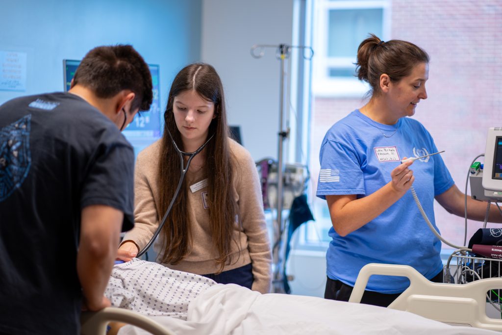 Three people are in a hospital room. One person listens to a patients chest with a stethoscope, another stands nearby using medical equipment, and a third watches attentively, all focused on patient care.