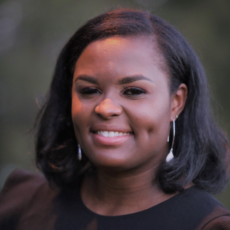 A woman with shoulder-length black hair, wearing a black top and silver hoop earrings, smiles warmly at the camera against a blurred outdoor background.