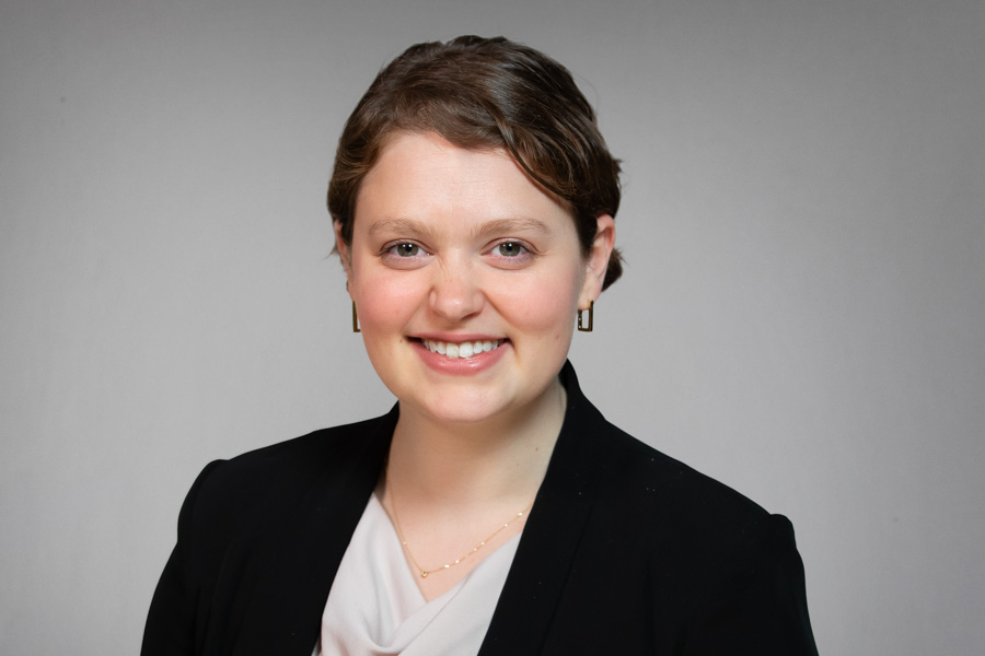 A person with short brown hair, wearing a black blazer over a light blouse, small hoop earrings, and a delicate necklace, smiles at the camera against a plain light gray background.