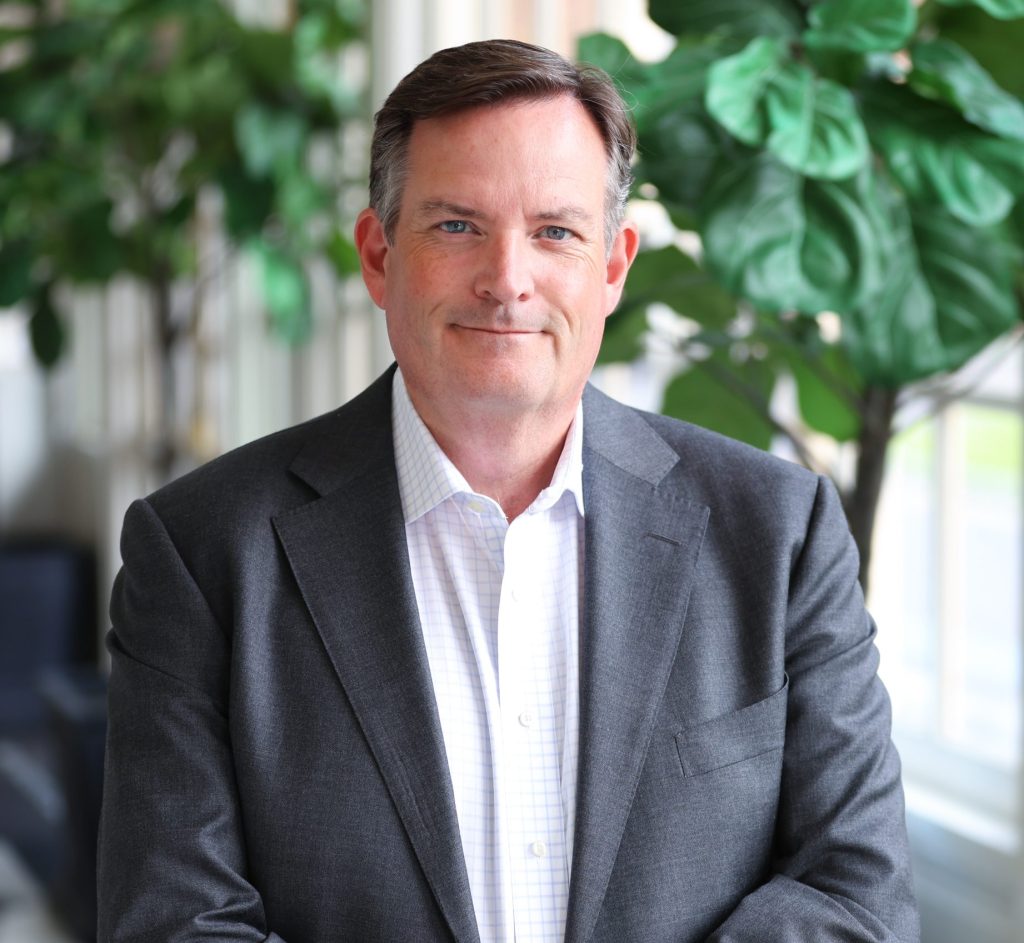 Brent Small, senior associate dean for research, smiles for a professional headshot wearing a gray suit jacket and white shirt indoors with large green plants and windows in the background.