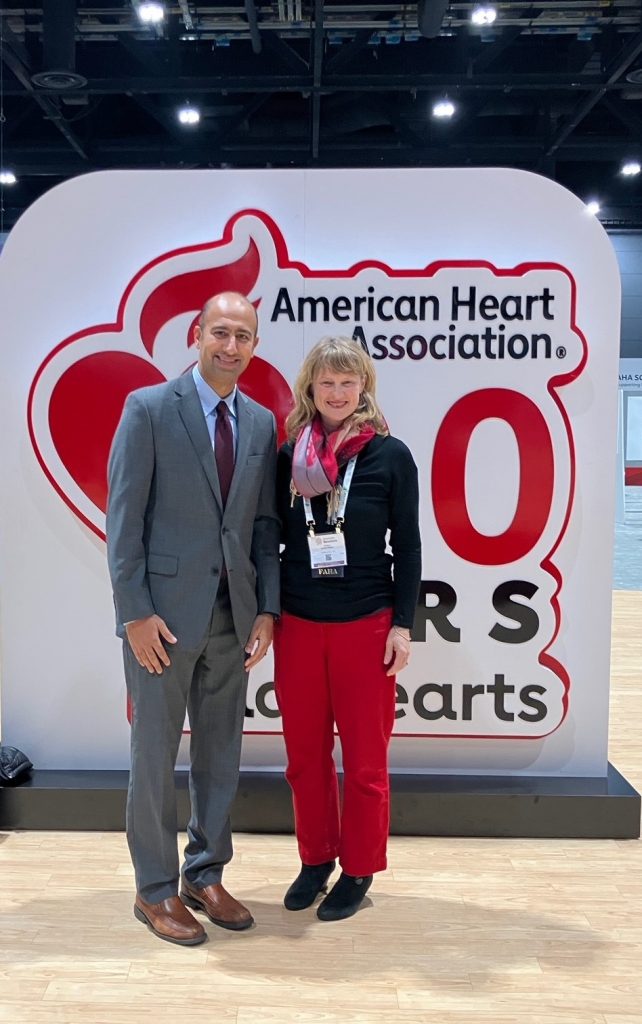Two people stand smiling in front of a large American Heart Association sign that reads 100 Years at an indoor event or conference. Both are wearing conference badges.