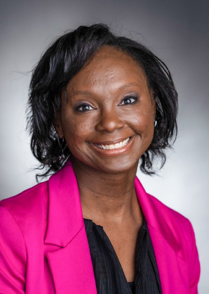 A woman with dark hair smiles at the camera. She is wearing a bright pink blazer over a black top, with a neutral gray background behind her.