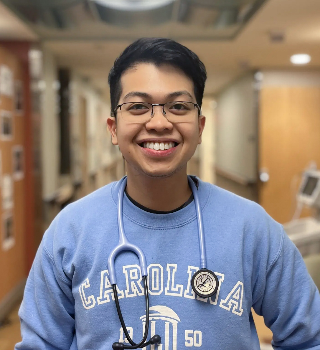 A smiling person wearing glasses, a blue Carolina sweatshirt, and a stethoscope stands in a brightly lit hallway, possibly in a hospital or clinic.