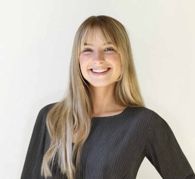 A young woman with long, straight blonde hair and bangs smiles warmly at the camera. She is wearing a black, vertically striped top and stands against a plain, light-colored background.