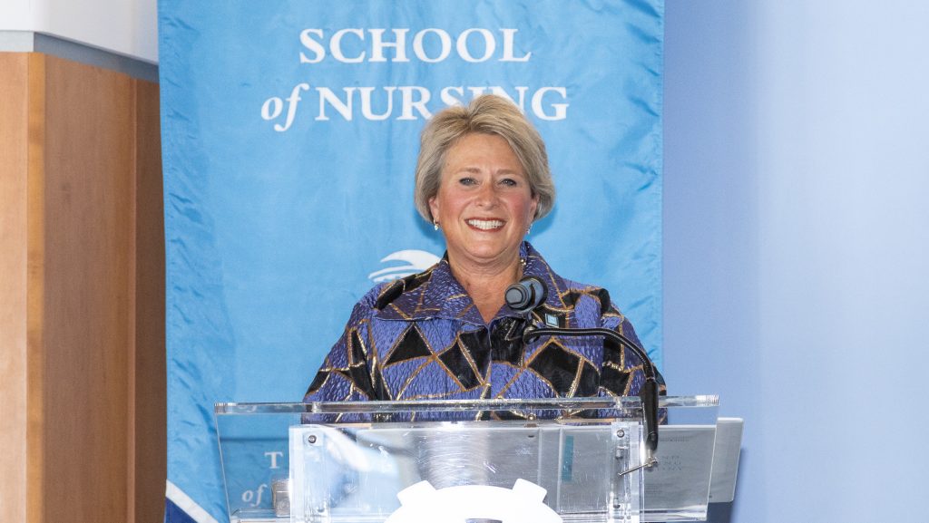 A woman stands at a podium, smiling, in front of a blue banner that reads School of Nursing. She is wearing a patterned jacket and speaking into a microphone.