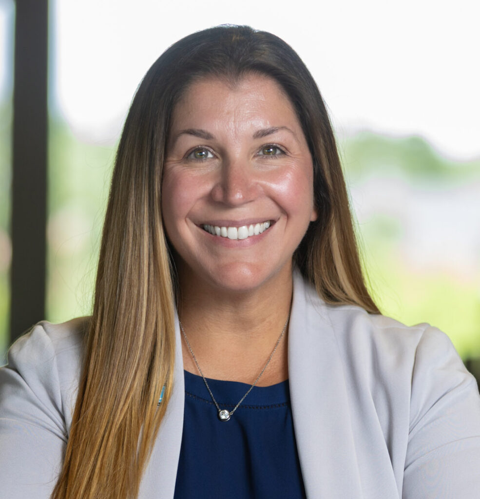 A woman with long brown hair wearing a light gray blazer and navy blue top sits at a table, smiling, with her arms folded. The background shows blurred windows and greenery.