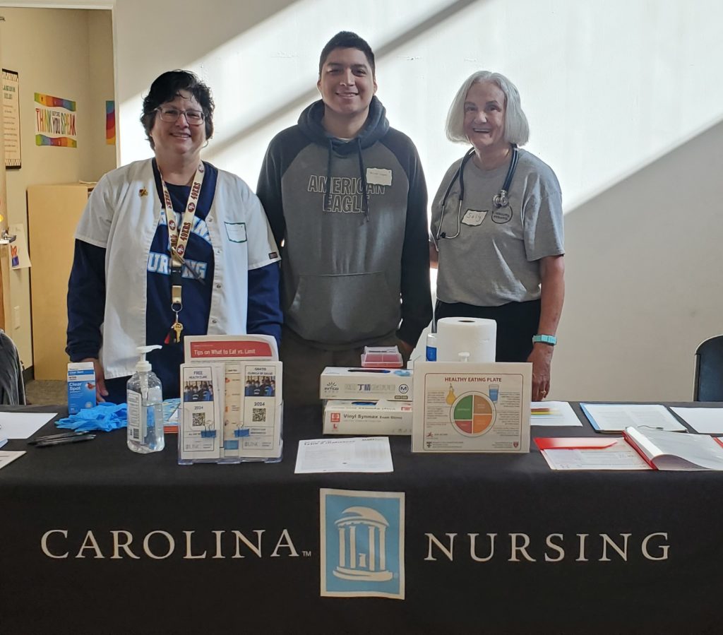 Three people stand behind a table labeled Carolina Nursing with brochures, sanitizer, and health materials displayed. Two women wear nursing attire, and a young man stands between them, all smiling at the camera.