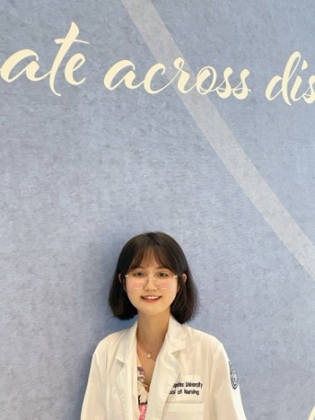 A young woman with short dark hair and glasses, wearing a white lab coat, stands smiling in front of a blue wall with cursive white text partially visible at the top.