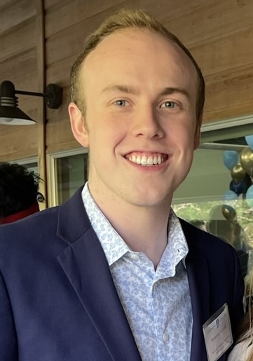 A young man in a blue suit jacket and patterned shirt smiles at the camera. He is indoors, and festive balloons are visible in the background. He wears a name tag on his jacket.