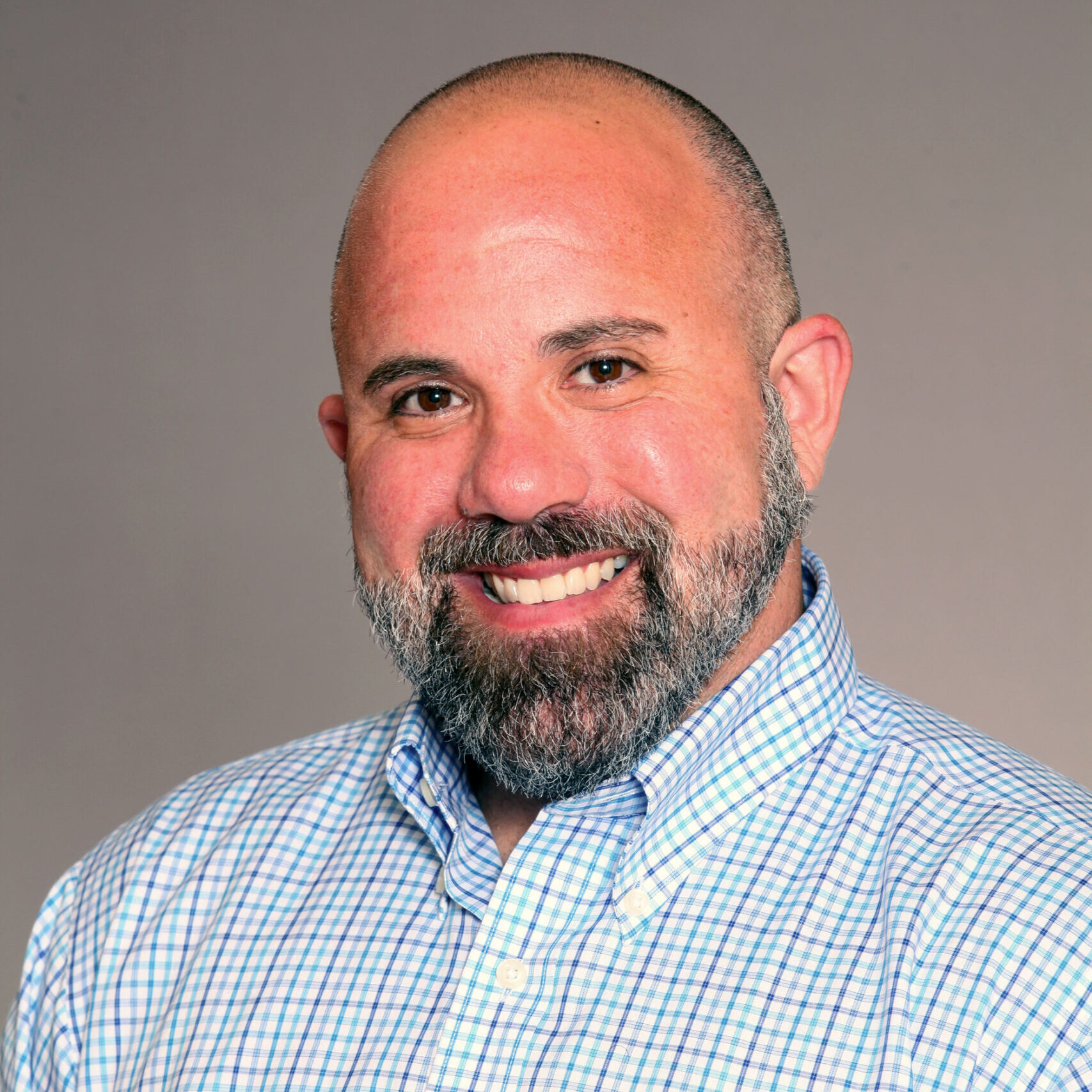 A smiling man with a bald head and a gray-and-black beard wearing a light blue and white checkered button-down shirt, posed in front of a plain, neutral background.
