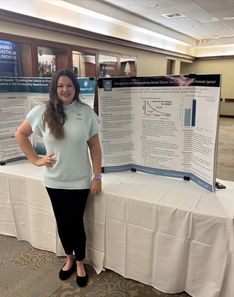 A woman stands next to a science poster presentation about mental illness in a conference room, smiling at the camera. She wears a light blue sweater and black pants; other posters and tables are visible in the background.