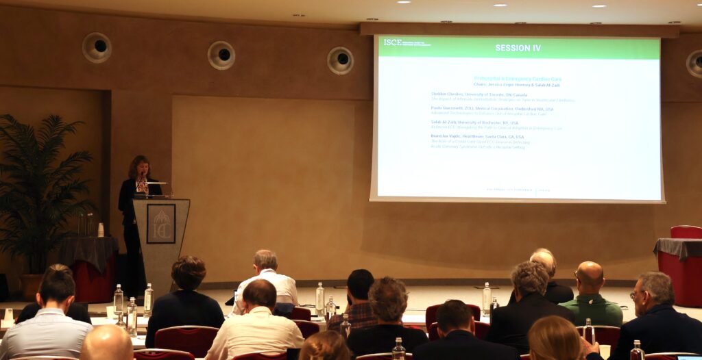 A speaker stands at a podium presenting to an audience in a conference room. A large screen behind her displays session information and a list of presenters. Attendees sit facing the stage, listening and taking notes.