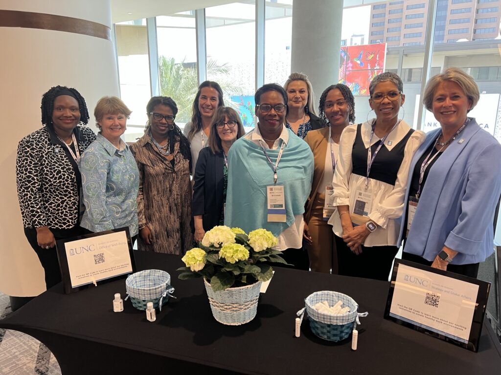 A group of eleven women stand smiling behind a table with flowers, baskets, and signs at a UNC event in a bright conference space with large windows.
