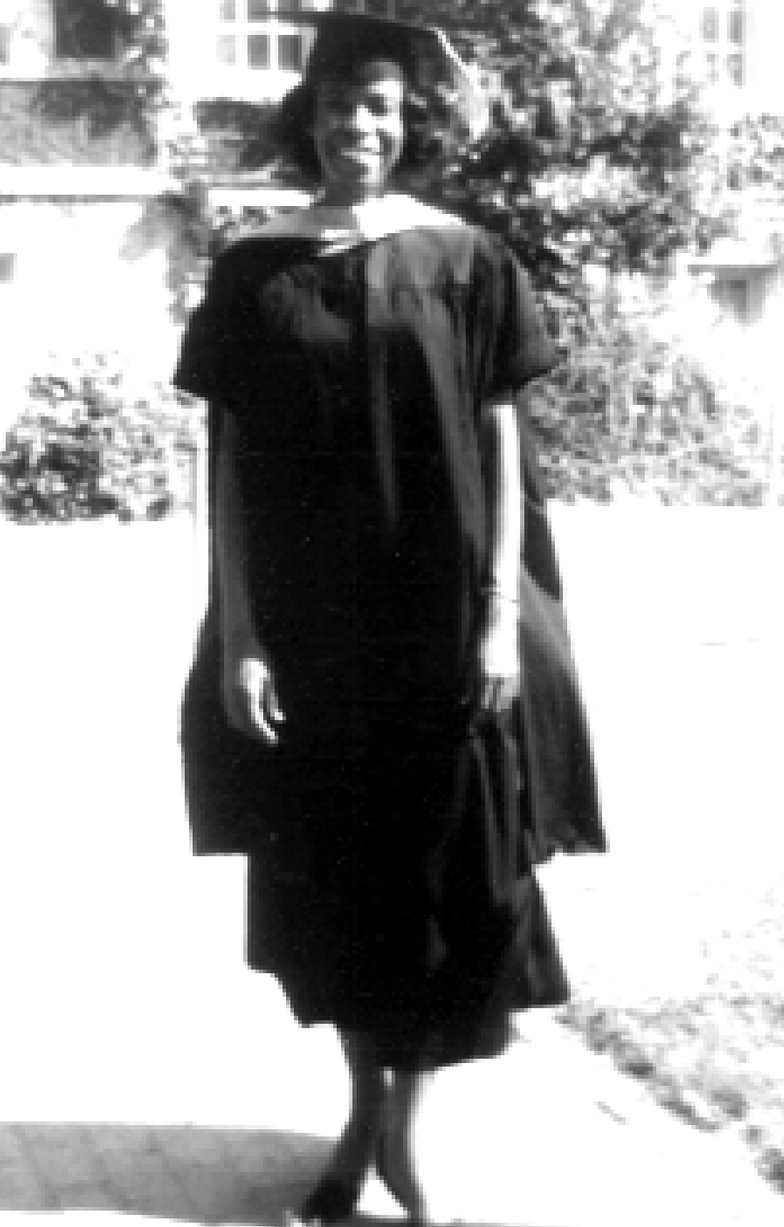 A person wearing a graduation cap and gown stands outside on a sunny day, smiling at the camera. Bushes and a building are visible in the background.