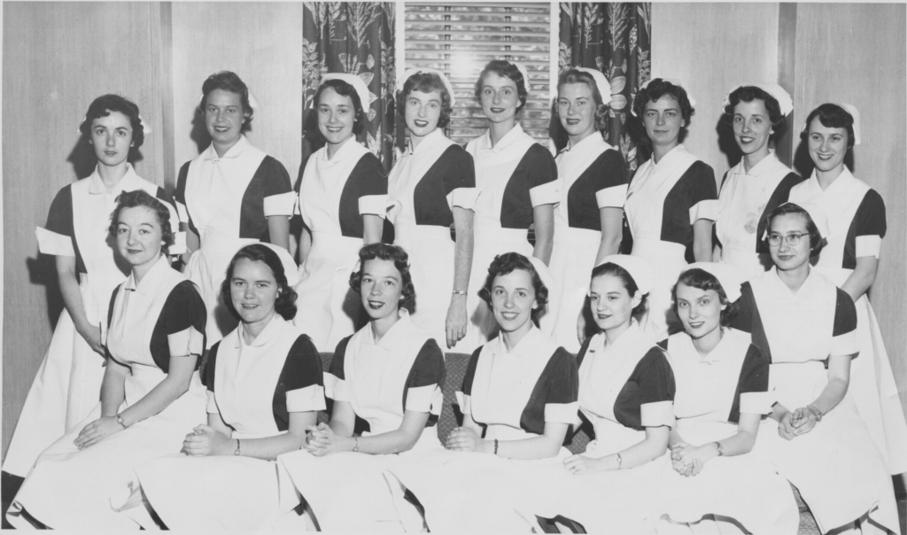 A group of fifteen women in matching nurse uniforms pose indoors in two rows, with some seated in front and others standing behind. They are smiling and looking at the camera.