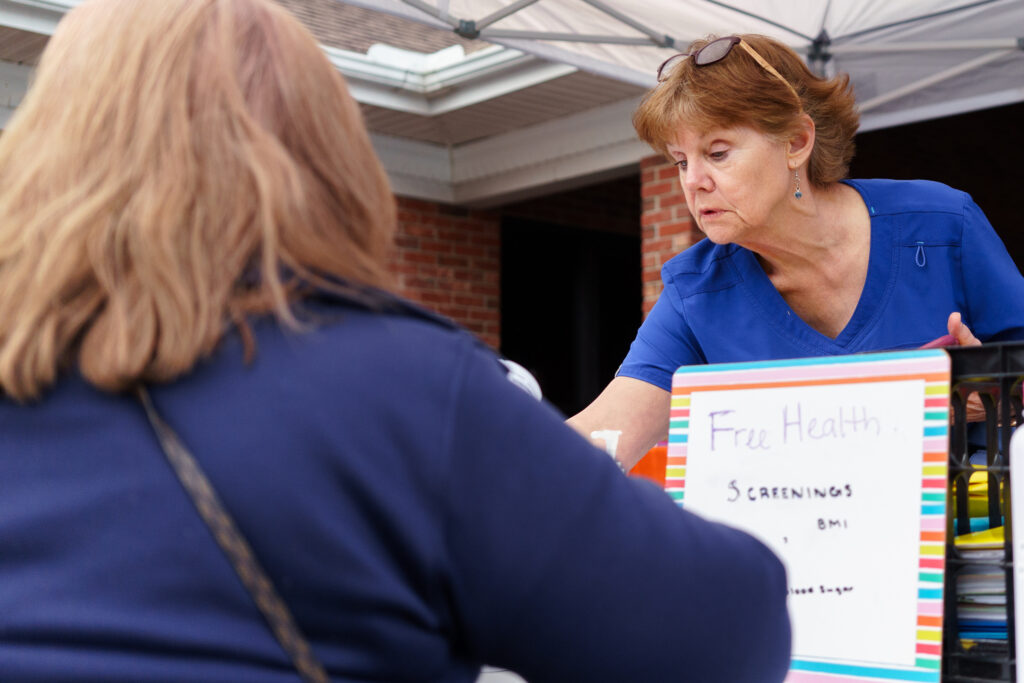 A woman in blue scrubs interacts with another woman at an outdoor health booth. A sign in the foreground reads Free Health Screenings BMI. They are under a white canopy.
