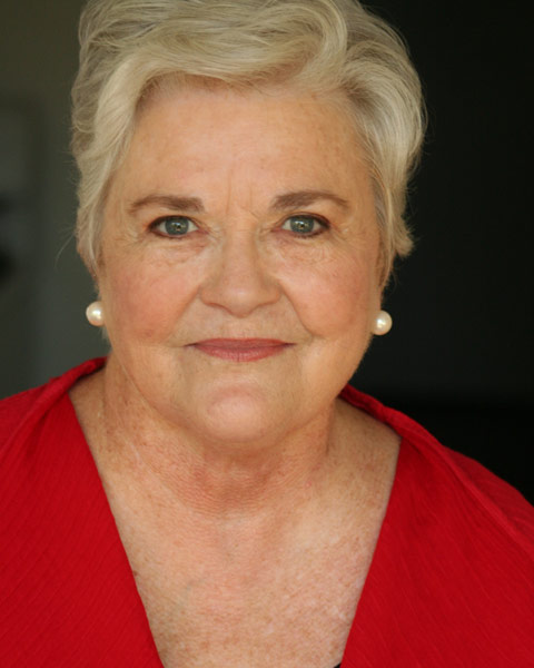 An older woman with short, gray hair and pearl earrings smiles gently. She is wearing a red top and is photographed against a dark background.