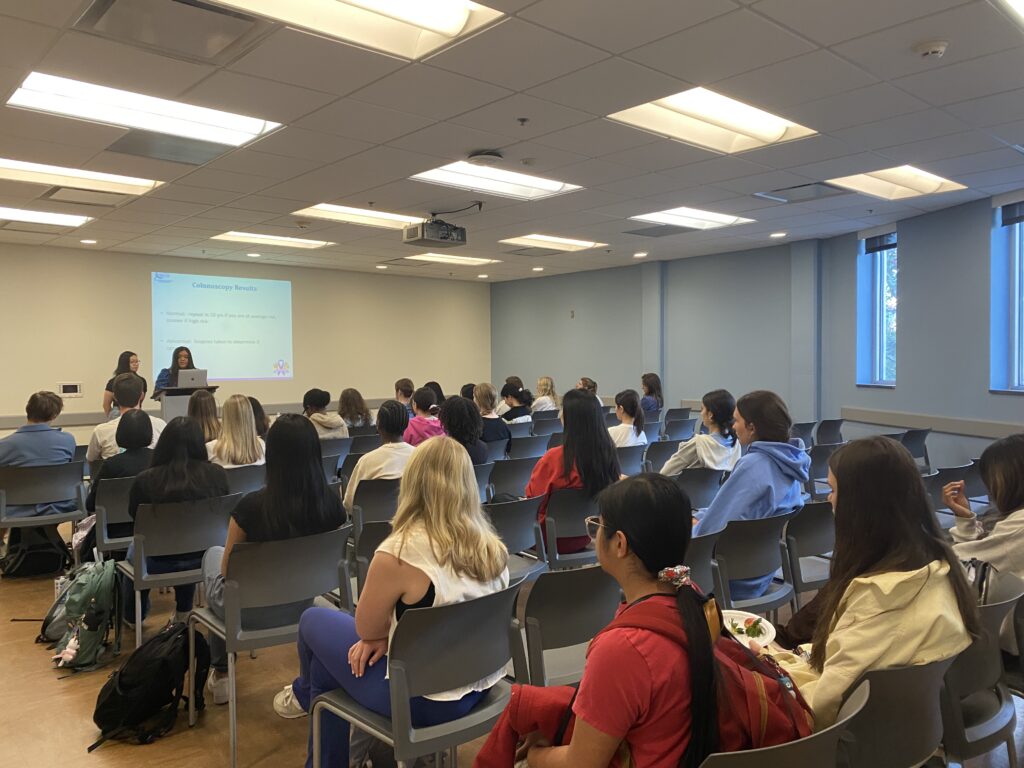 A group of students sits in a classroom facing two presenters at the front, where a presentation is projected onto a screen. The room has rows of gray chairs and large windows on one side.