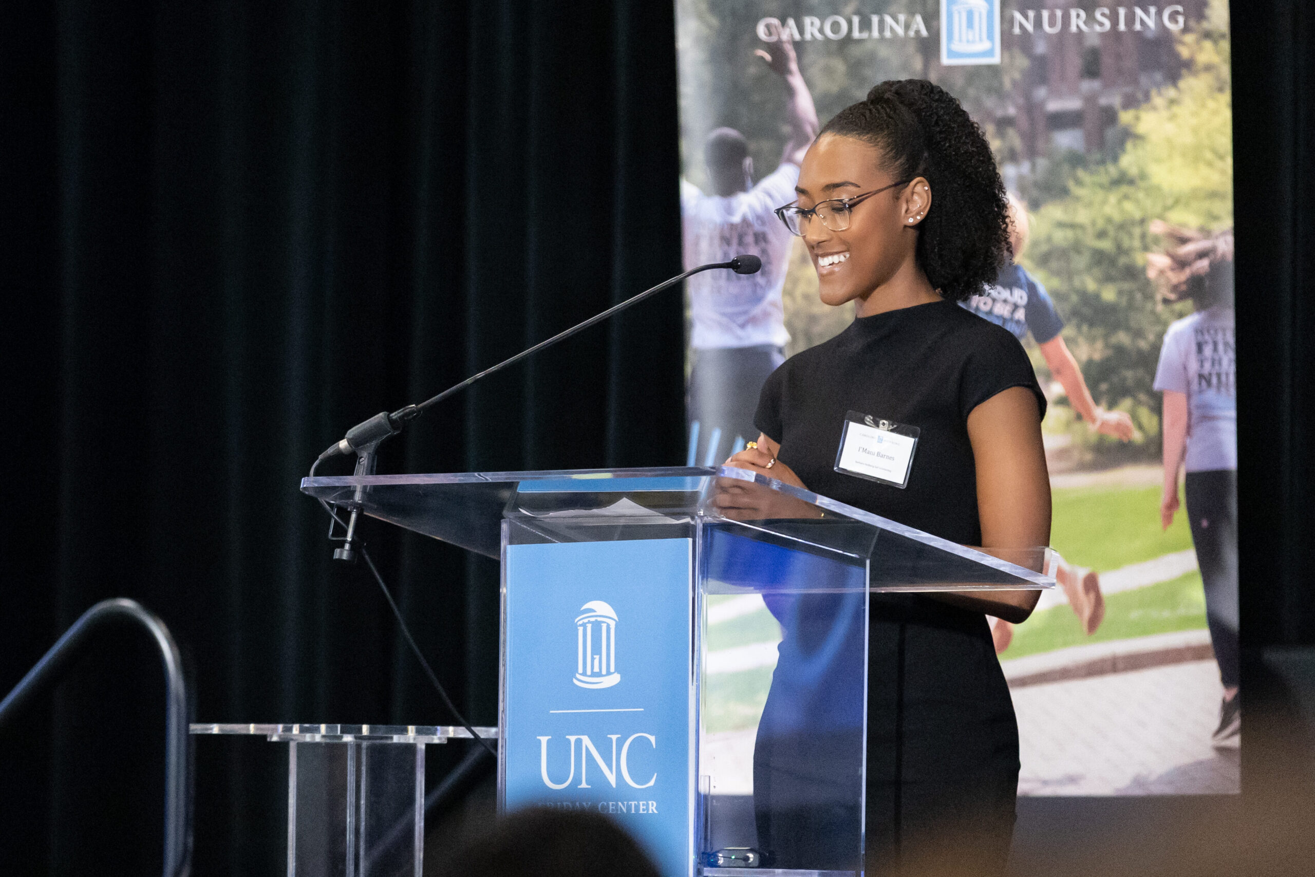 A woman stands at a clear podium with a UNC sign, smiling as she speaks into a microphone. A banner reading Carolina Nursing is visible in the background.