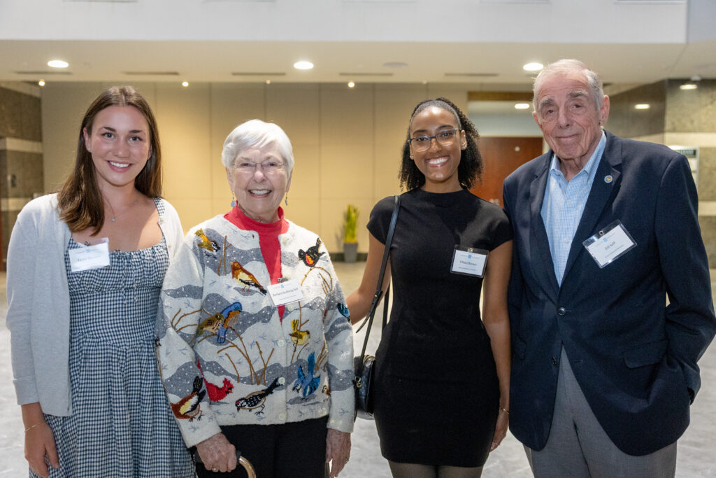 Four people stand together indoors, smiling at the camera. Two women on the left wear a checked dress and a sweater with birds, while the woman on the right wears a black dress and the man on the far right wears a dark suit.