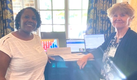 Two women stand smiling at a table displaying laptops and informational materials in front of a bright window, with patterned curtains in the background.