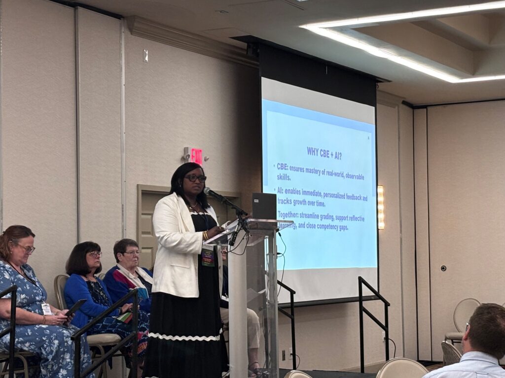A woman stands at a podium presenting to an audience in a conference room. Behind her, a projector screen displays a slide titled WHY CBE - AI? with bulleted points. Four seated women are visible to the left.