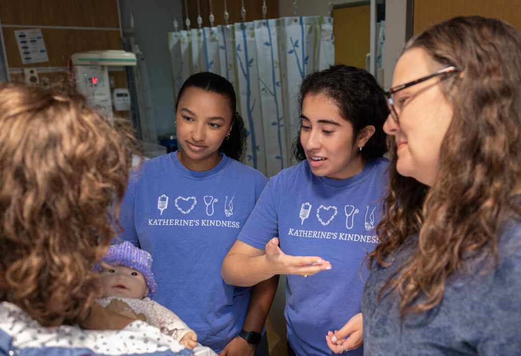 Four women, three wearing blue Katherines Kindness shirts, stand together in a hospital room. One woman holds a baby doll dressed in a purple hat while the others interact and smile warmly.