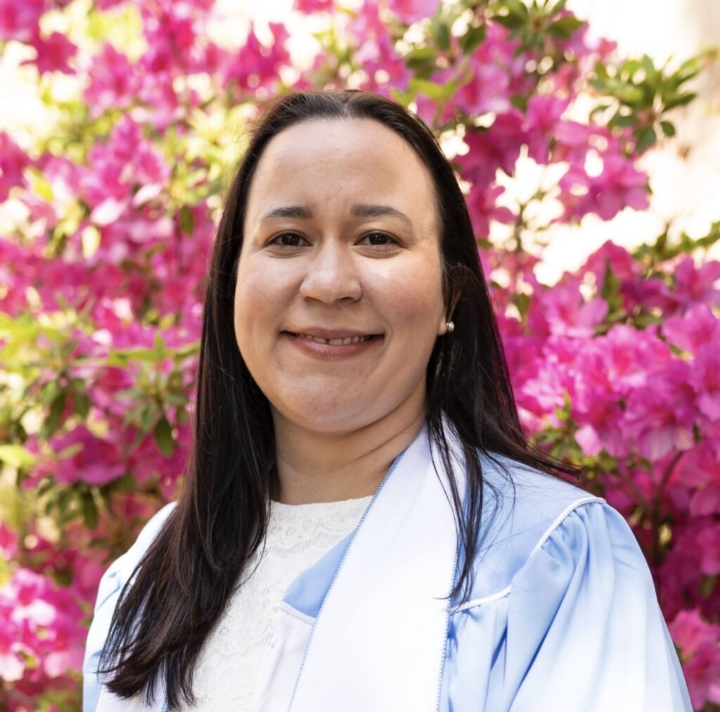 A person with long dark hair, wearing a light blue graduation gown and white top, smiles in front of blooming pink flowers on a sunny day.