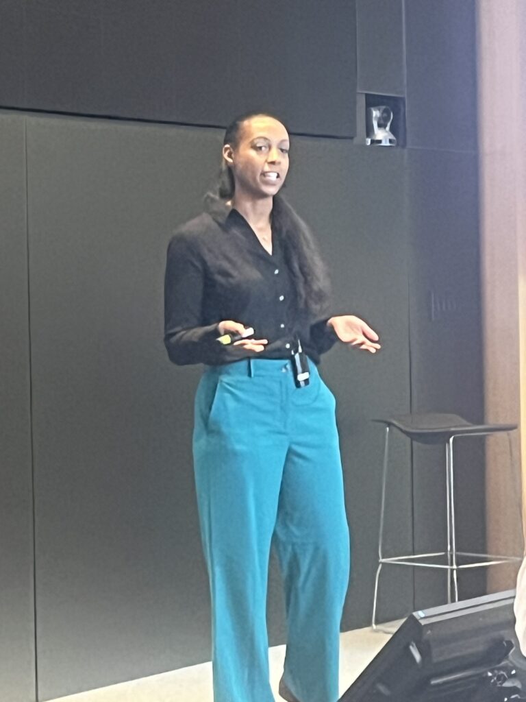 A woman, Kandyce Brennan, with long hair in a ponytail, wearing a black shirt and teal pants, is standing and speaking in front of a dark wall, gesturing with her hands. A black stool is visible in the background.