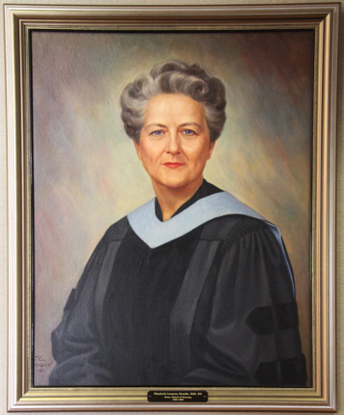 A painted portrait of an older woman with gray, styled hair, wearing a black academic gown with light blue accents, set against a soft, multicolored background and framed in a gold frame.