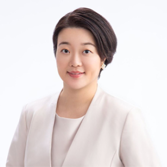 A woman with short dark hair, wearing a light-colored blazer and blouse, smiles slightly while facing the camera against a plain white background.