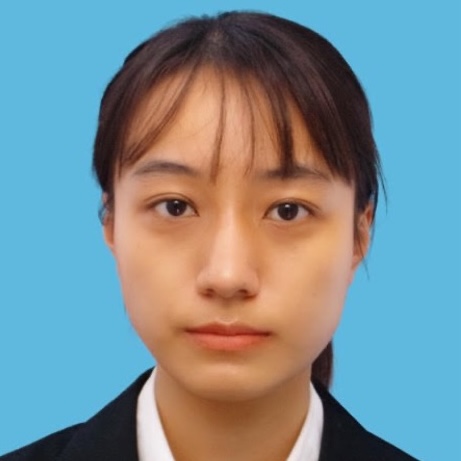 A young woman with straight brown hair, wearing a black suit jacket and white collared shirt, facing forward against a solid blue background.