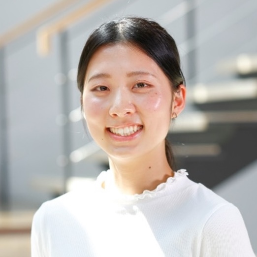 A young woman with long dark hair tied back, wearing a white top, smiles at the camera. The background is softly blurred, showing stairs and railings in a sunlit indoor setting.
