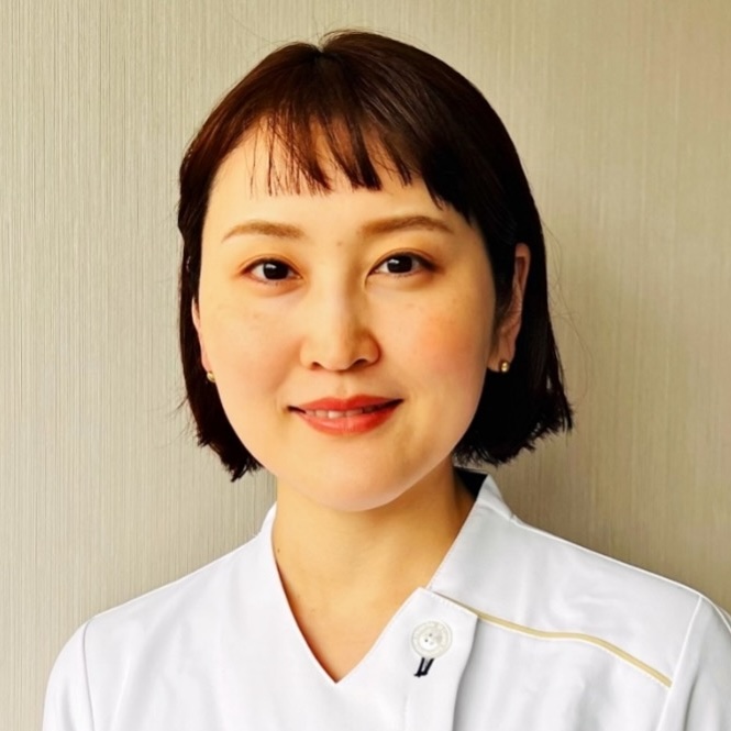 A woman with short dark hair and bangs, wearing a white collared uniform, stands in front of a light-colored wall and smiles gently at the camera.