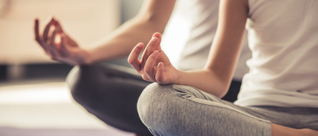 Two people sitting cross-legged on yoga mats, wearing light-colored tops and gray leggings, with hands resting on knees in a meditation pose. The focus is on their hands and lower bodies.