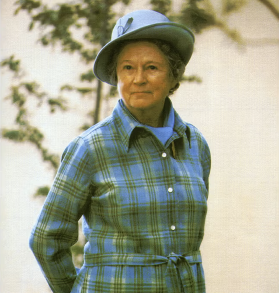 An older woman stands outdoors, wearing a blue plaid coat with a belt and a matching blue hat. She looks slightly to the side, with trees and a light-colored background behind her.