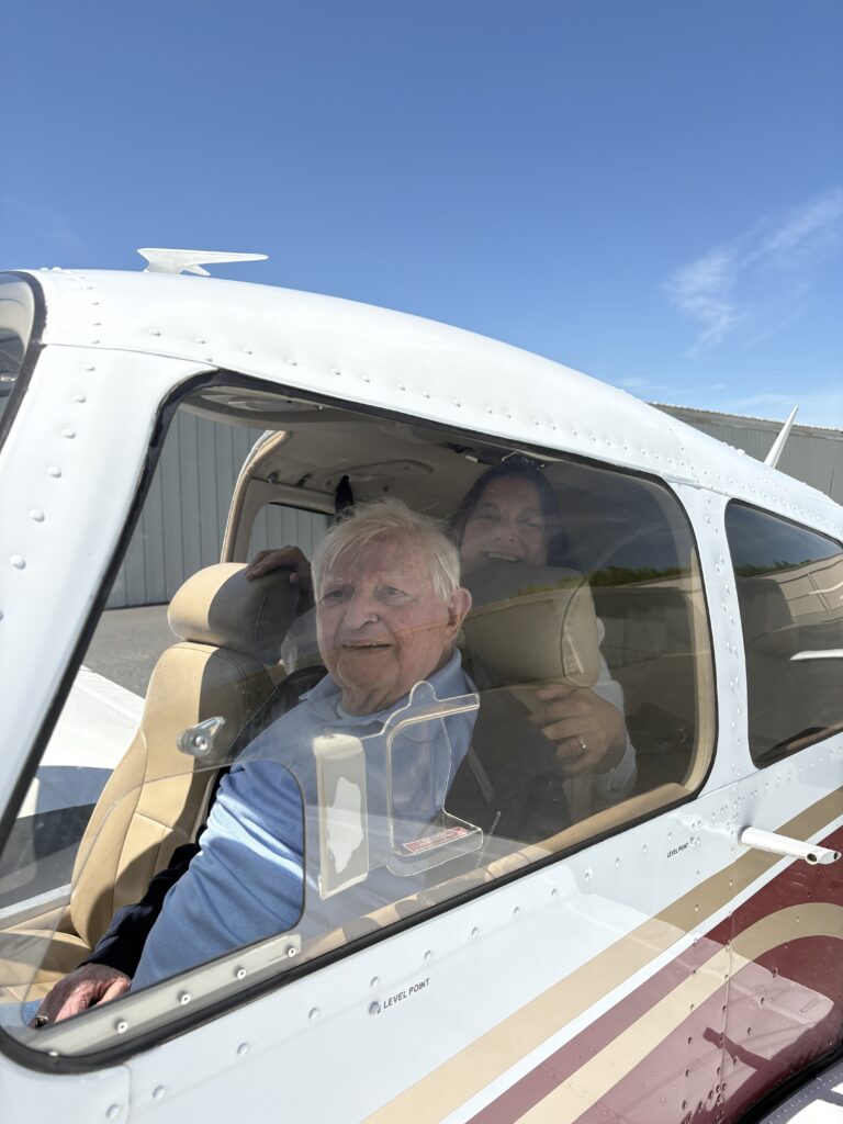 Two people sit inside a small airplane on a sunny day; the older man in front smiles, while another person sits behind him. The aircraft is parked on the ground near a hangar.