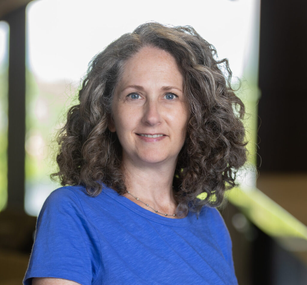 A woman with curly brown hair wearing a blue shirt stands indoors, smiling slightly. The background is softly blurred, with large windows letting in natural light.
